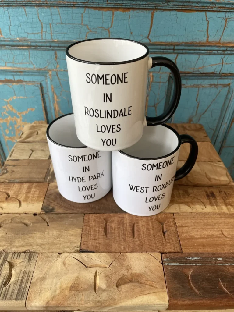 Three white mugs with black handles display messages about someone loving you in Roslindale, Hyde Park, and West Roxbury.
